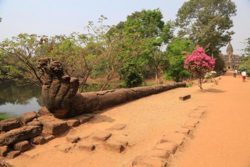  An ancient stone sculpture of a serpent or dragon beside a dirt path leading to a distant temple, with people walking amidst lush greenery and a blooming pink tree.