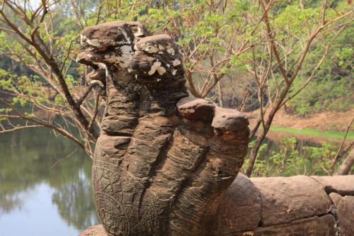 A weathered stone statue, possibly a serpent or dragon, in an outdoor setting near a body of water, surrounded by trees and vegetation.
